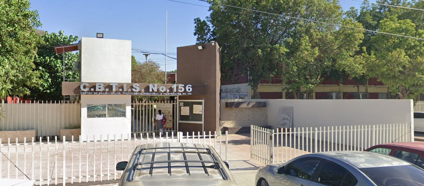 Fotografía en la que se muestra la entrada del plantel, conformada por una reja de color blanco que rodea las instalaciones. Al fondo se encuentra una reja blanca que da acceso al plantel y sobre ella, hay una placa de color café que en letras grandes grises dicen Cebetis NÚMERO 156.